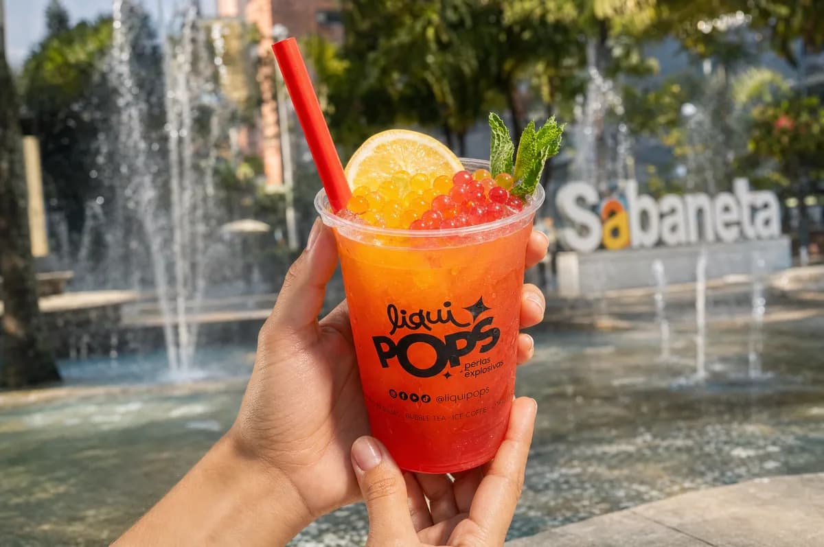 Manos sosteniendo una limonada roja con perlas explosivas LiquiPops amarillas y rojas y rodaja de limon, frente a la fuente de Sabaneta