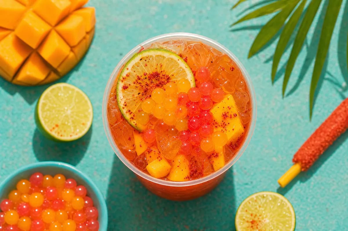 Mangonada de mango con chamoy, trozos de mango, lima y perlas explosivas rojas y amarillas LiquiPops — vista superior sobre fondo turquesa