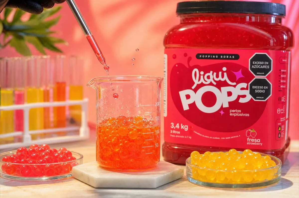 Vaso de precipitado con perlas explosivas naranjas siendo goteadas con pipeta, junto a tubos de ensayo de colores, bowls de perlas rojas y amarillas y el tarro LiquiPops sabor fresa — control de calidad de perlas explosivas