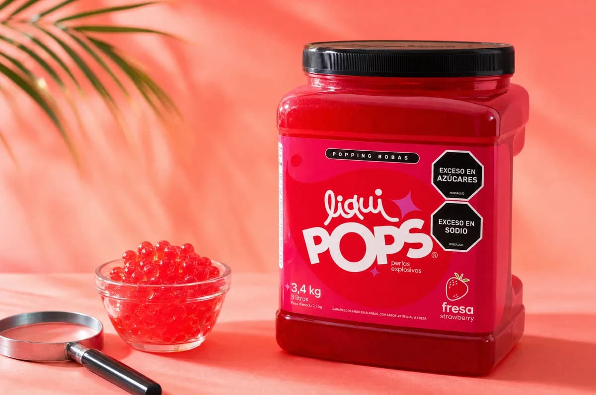 Tarro de perlas explosivas LiquiPops sabor fresa de 3,4 kg junto a un bowl con perlas rojas y una lupa sobre fondo coral con hoja de palma