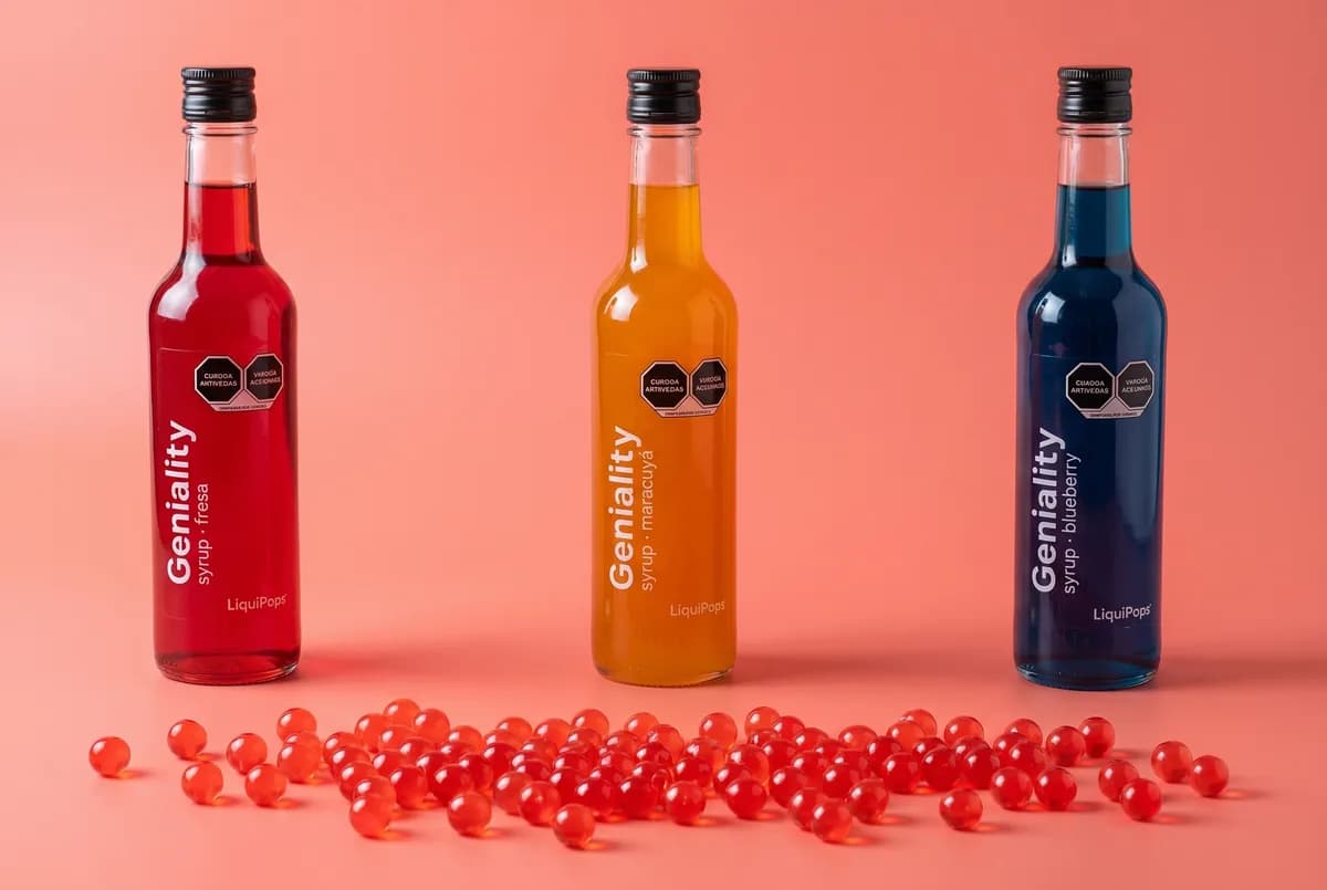 Tres siropes Geniality LiquiPops sabores fresa, maracuyá y blueberry alineados con perlas explosivas rojas regadas sobre fondo coral — bodegón de producto