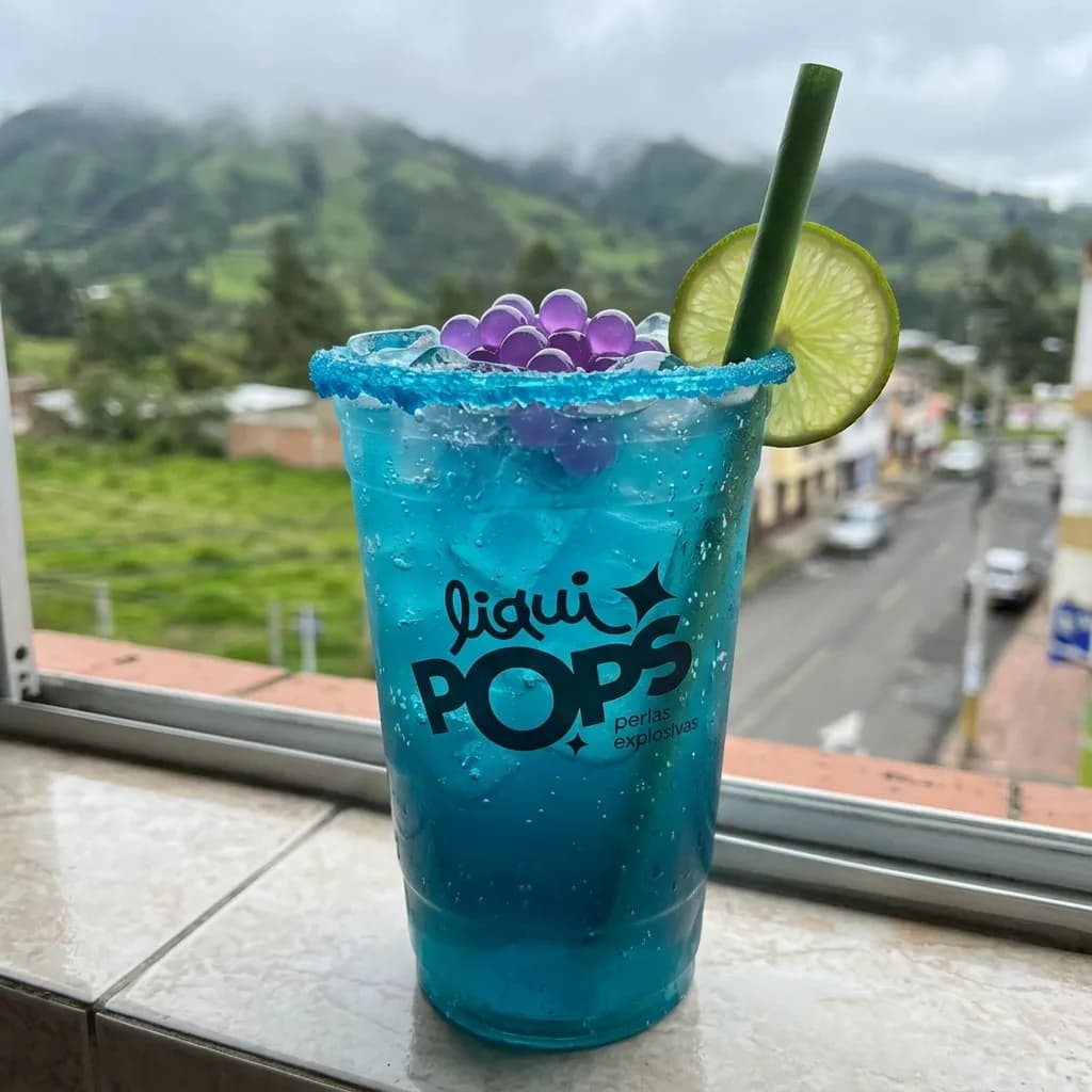 Soda azul con perlas explosivas moradas LiquiPops, borde escarchado y limon, junto a una ventana con paisaje montanoso de los Andes