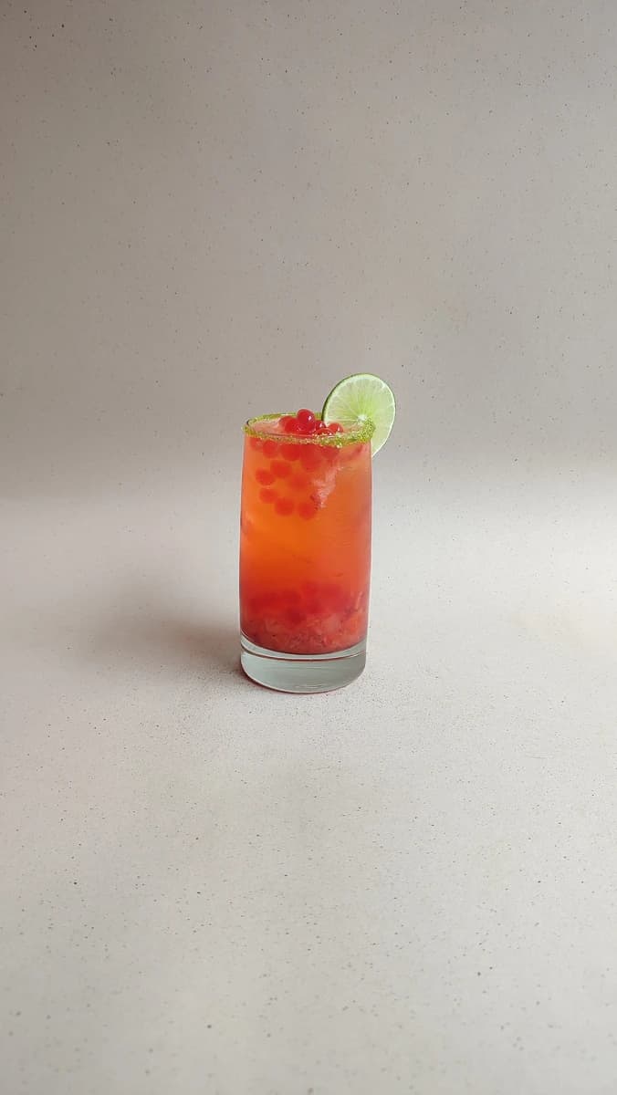 Cóctel de fresa y aguardiente con perlas explosivas LiquiPops, borde escarchado y limón en vaso alto sobre fondo gris