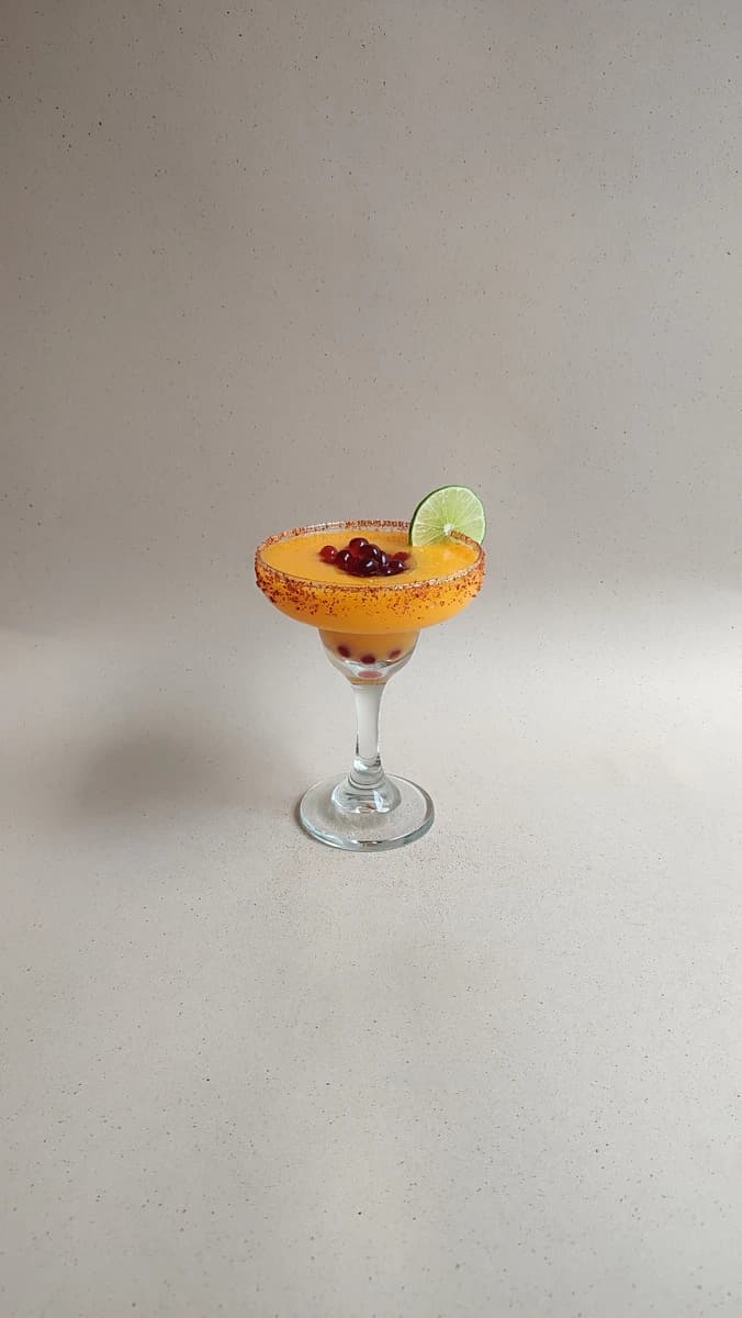 Margarita de mango con perlas explosivas LiquiPops, escarchado de chile y rodaja de limón en copa sobre fondo gris