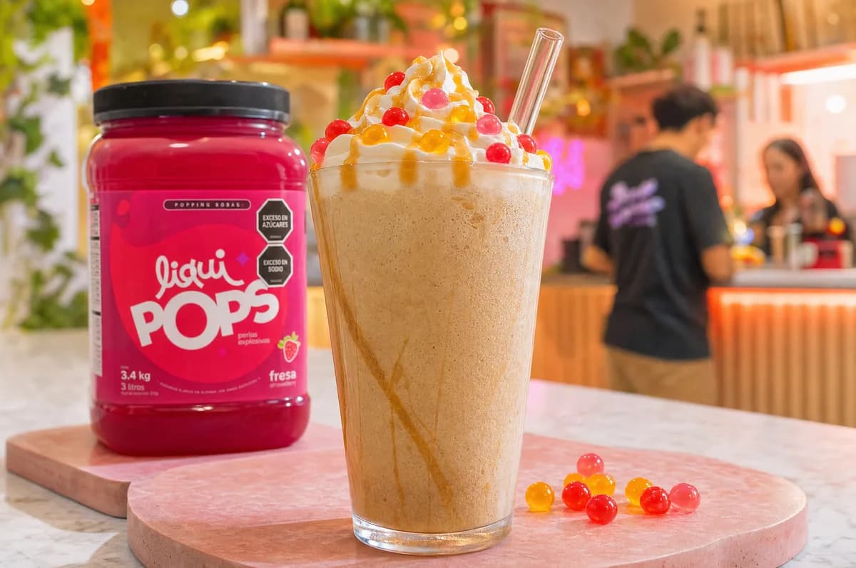 Frappé cremoso con crema batida y perlas explosivas LiquiPops junto al envase sabor fresa de la marca — barra de café con ambiente juvenil