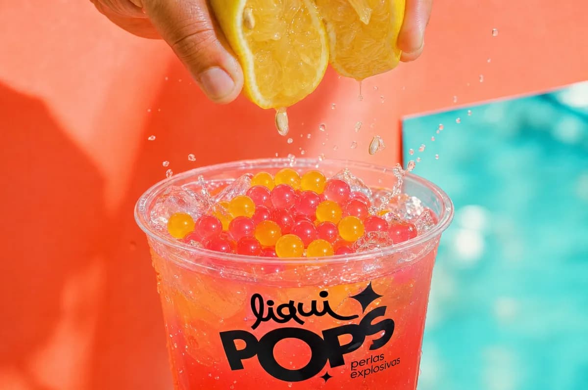 Manos exprimiendo limón sobre un vaso de limonada con perlas explosivas rojas y amarillas LiquiPops — fondo coral y turquesa