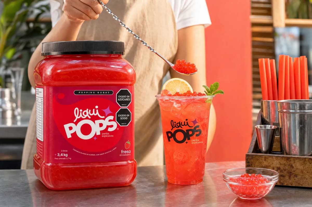 Persona sirviendo con cuchara perlas explosivas rojas de fresa sobre una limonada roja con rodaja de limón y menta en vaso LiquiPops, junto al tarro sabor fresa en la barra