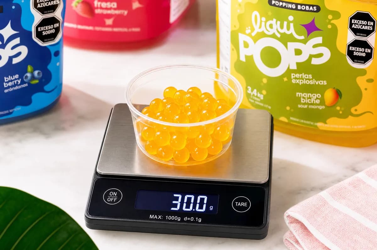 Porción de 30 gramos de perlas explosivas LiquiPops sabor mango biche sobre báscula digital junto a tarros de blueberry, fresa y mango