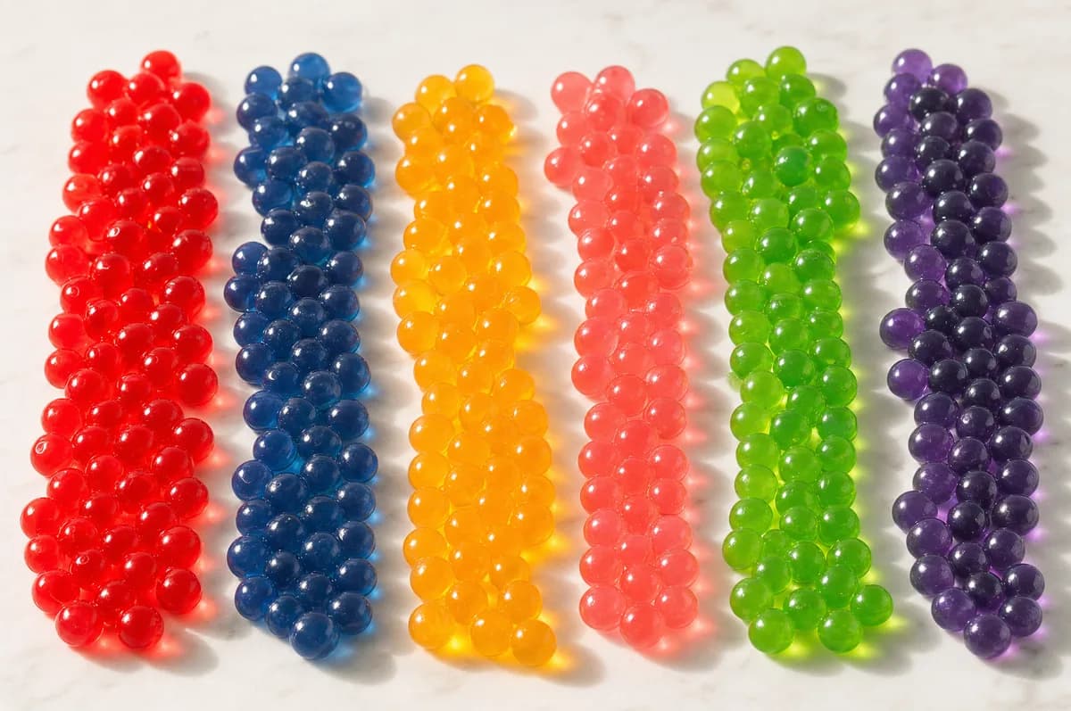 Perlas explosivas LiquiPops en seis colores ordenadas en filas sobre mármol — rojo, azul, naranja, rosa, verde y morado