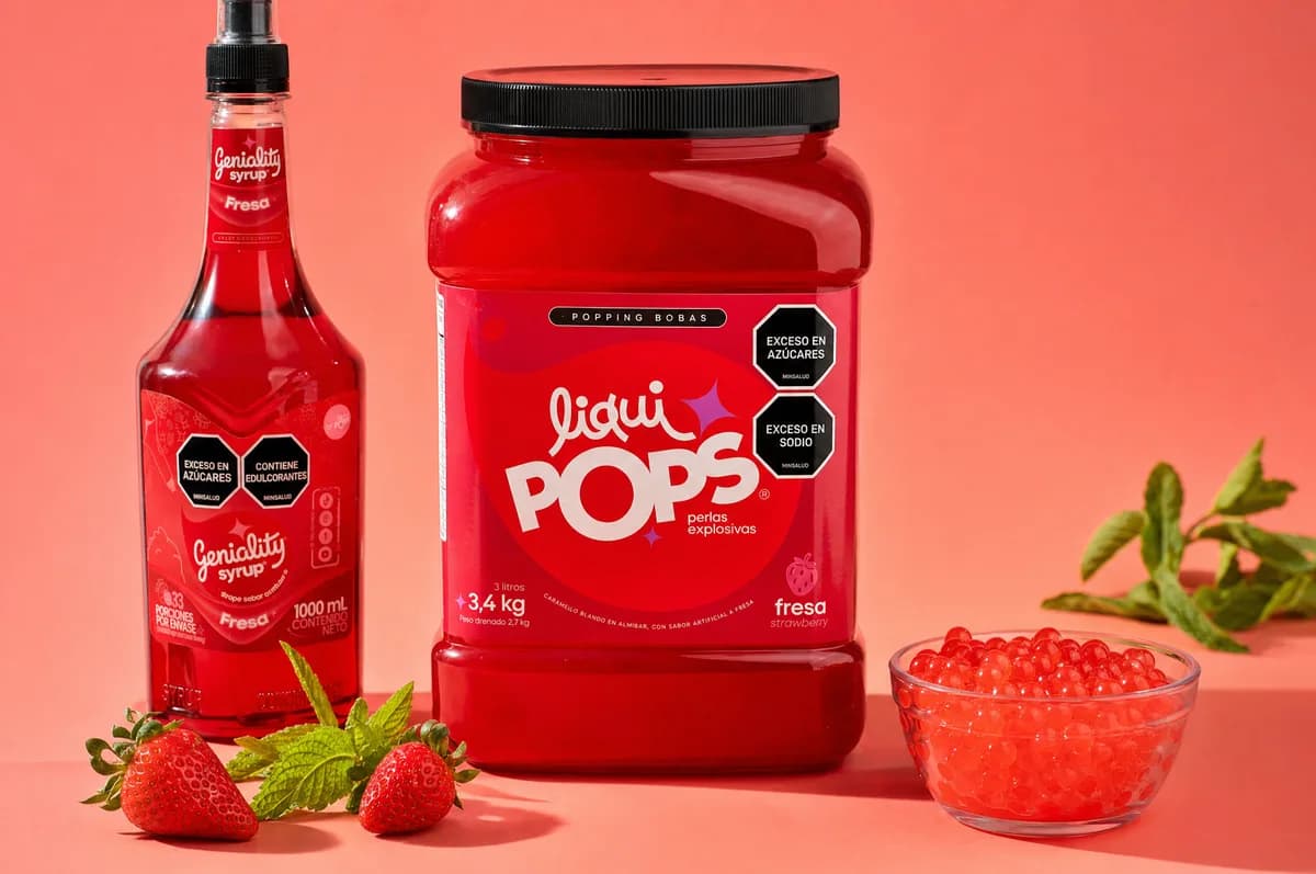 Producto LiquiPops: sirope Geniality sabor fresa, tarro de perlas explosivas fresa y bowl de perlas rojas con fresas frescas — bodegón sobre fondo coral