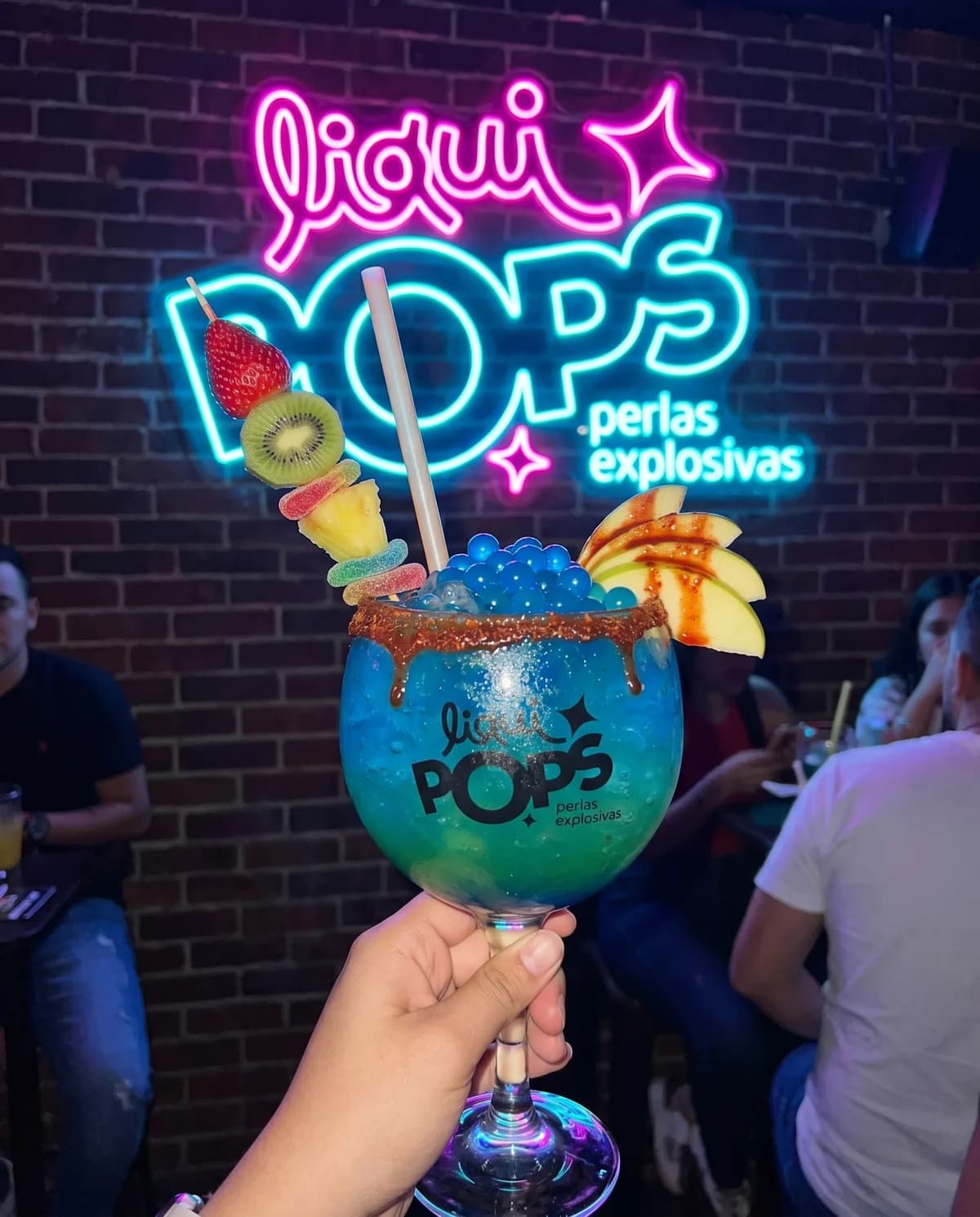 Mano sosteniendo una copa de soda loca con perlas explosivas y pasador de frutas LiquiPops en un bar bajo el letrero neón