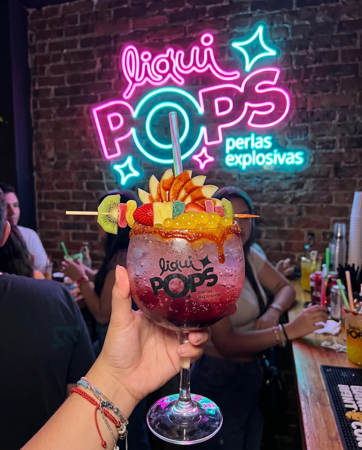Mano sosteniendo una soda loca de mango con perlas explosivas azules LiquiPops en un bar bajo el letrero neón — pasador de frutas y escarcha de chamoy