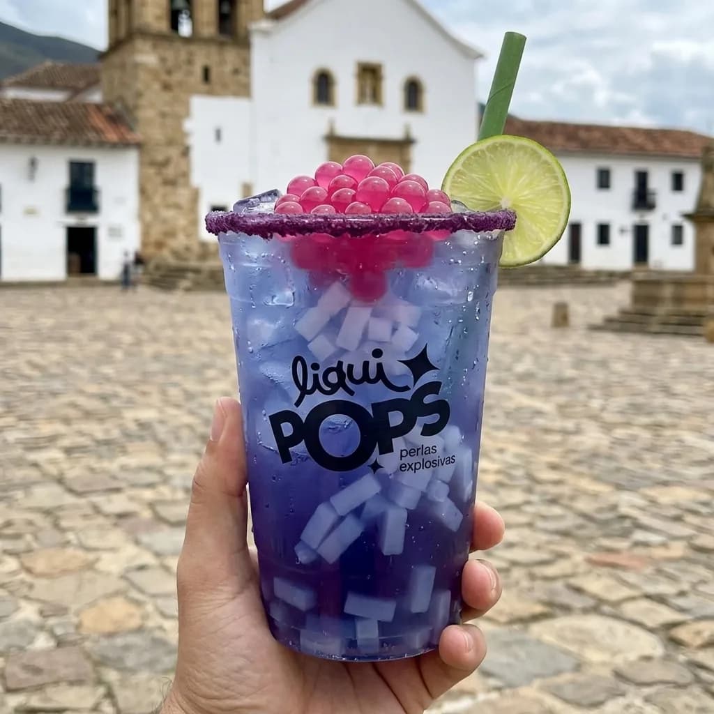 Mano sosteniendo una soda morada con perlas explosivas de fresa y cubos de coco LiquiPops, frente a una plaza colonial colombiana