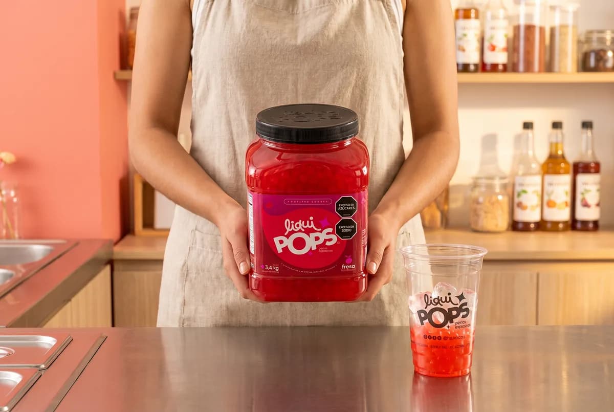 Persona sostiene un tarro de 3.4 kg de perlas explosivas de fresa LiquiPops junto a un vaso con perlas — perlas explosivas LiquiPops al por mayor