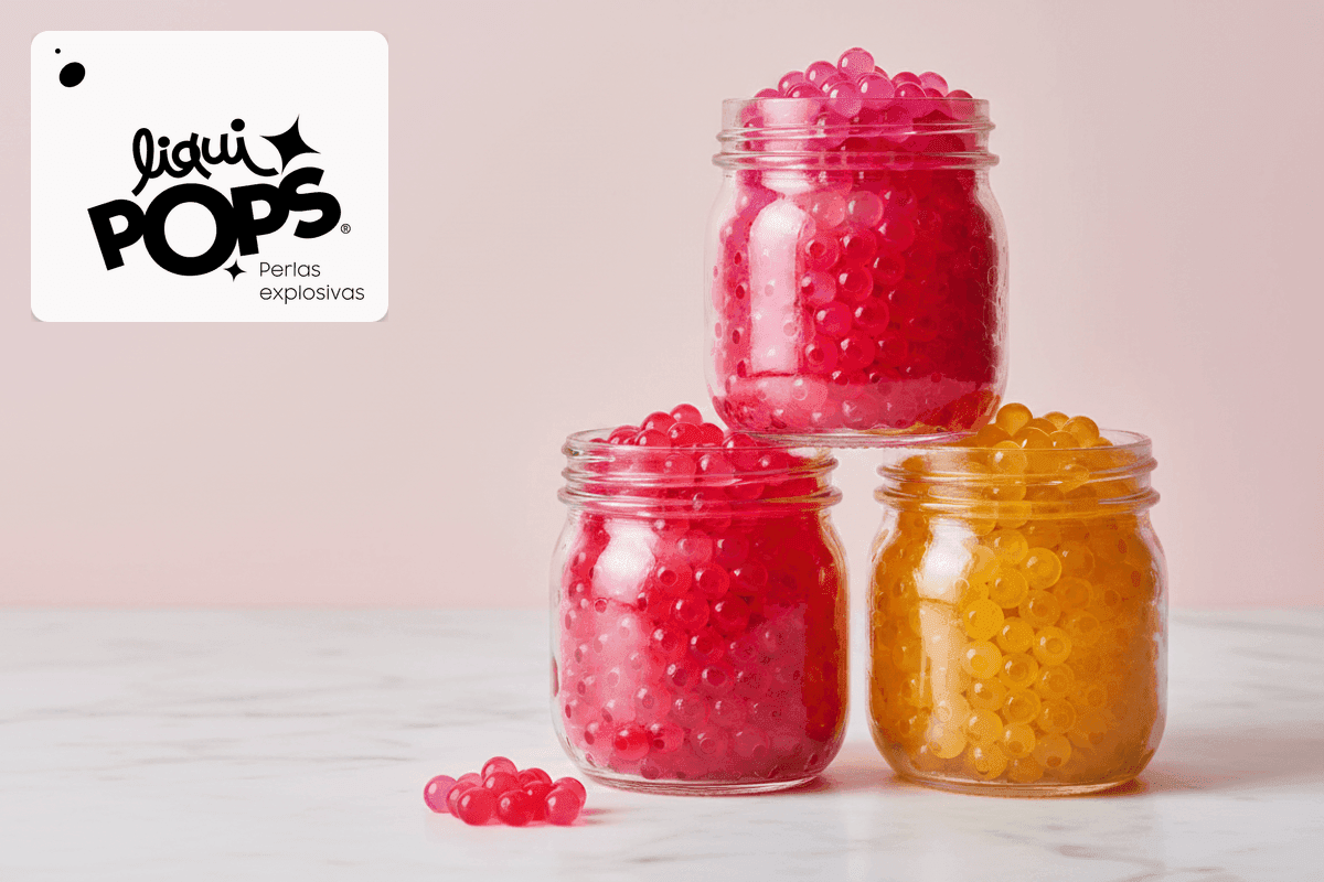 Tres frascos de perlas explosivas LiquiPops en pirámide: rosa, rojo y amarillo, sobre mármol blanco con pared rosa de fondo y perlas sueltas en el piso