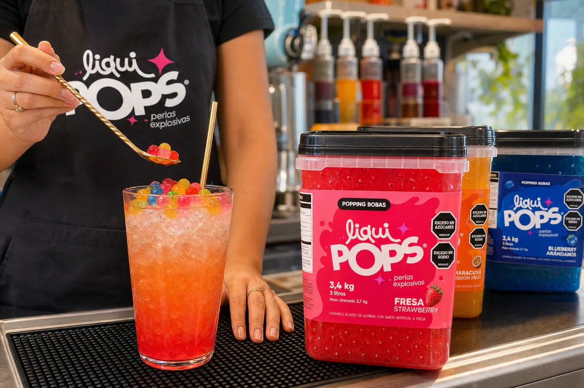 Persona con delantal LiquiPops sirviendo con cuchara perlas explosivas de colores sobre una bebida roja de fresa con hielo, junto a tarros de perlas LiquiPops sabor fresa, maracuyá y blueberry en la barra
