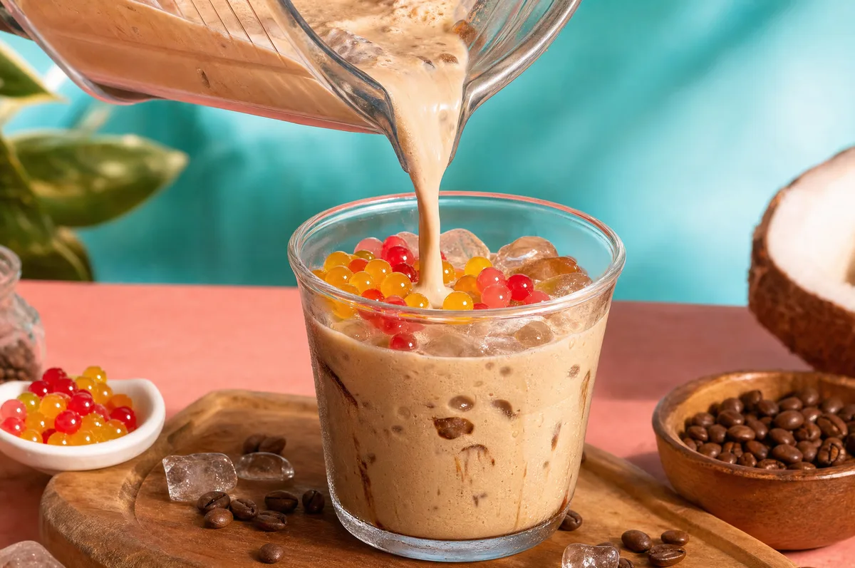 Café helado con leche de coco sirviéndose sobre hielo con perlas explosivas rojas y amarillas LiquiPops — granos de café y coco al lado