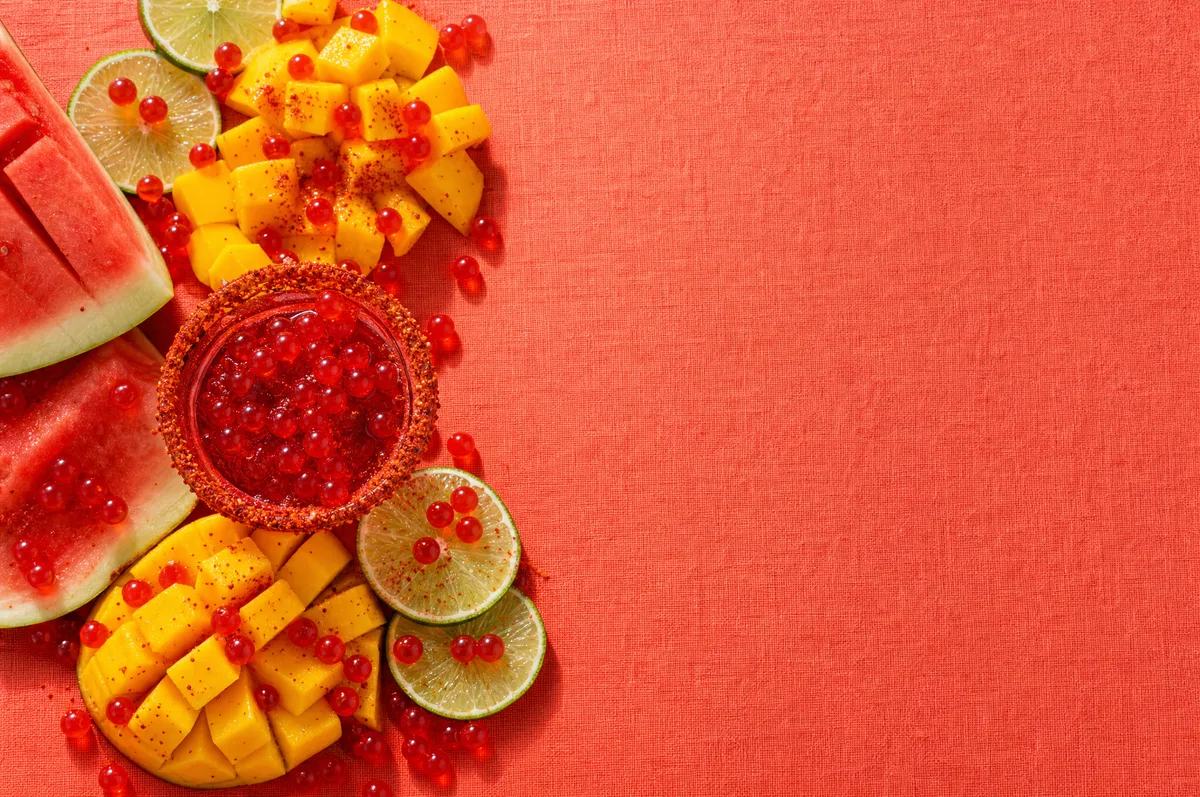 Flatlay de mango, sandia y limon con perlas explosivas rojas LiquiPops sobre fondo rojo, ingredientes para bebidas