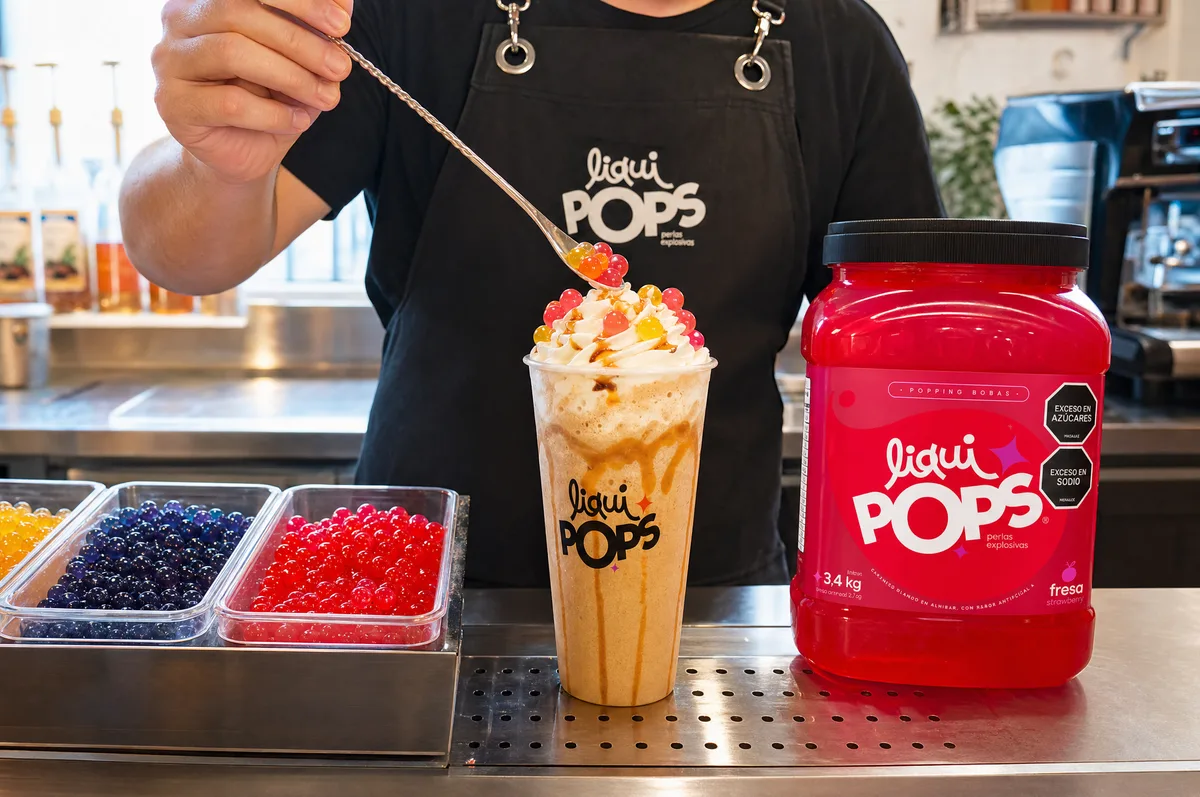 Barista sirviendo perlas explosivas sabor fresa LiquiPops sobre un frappé con crema batida en barra de cafetería — envase rojo de la marca