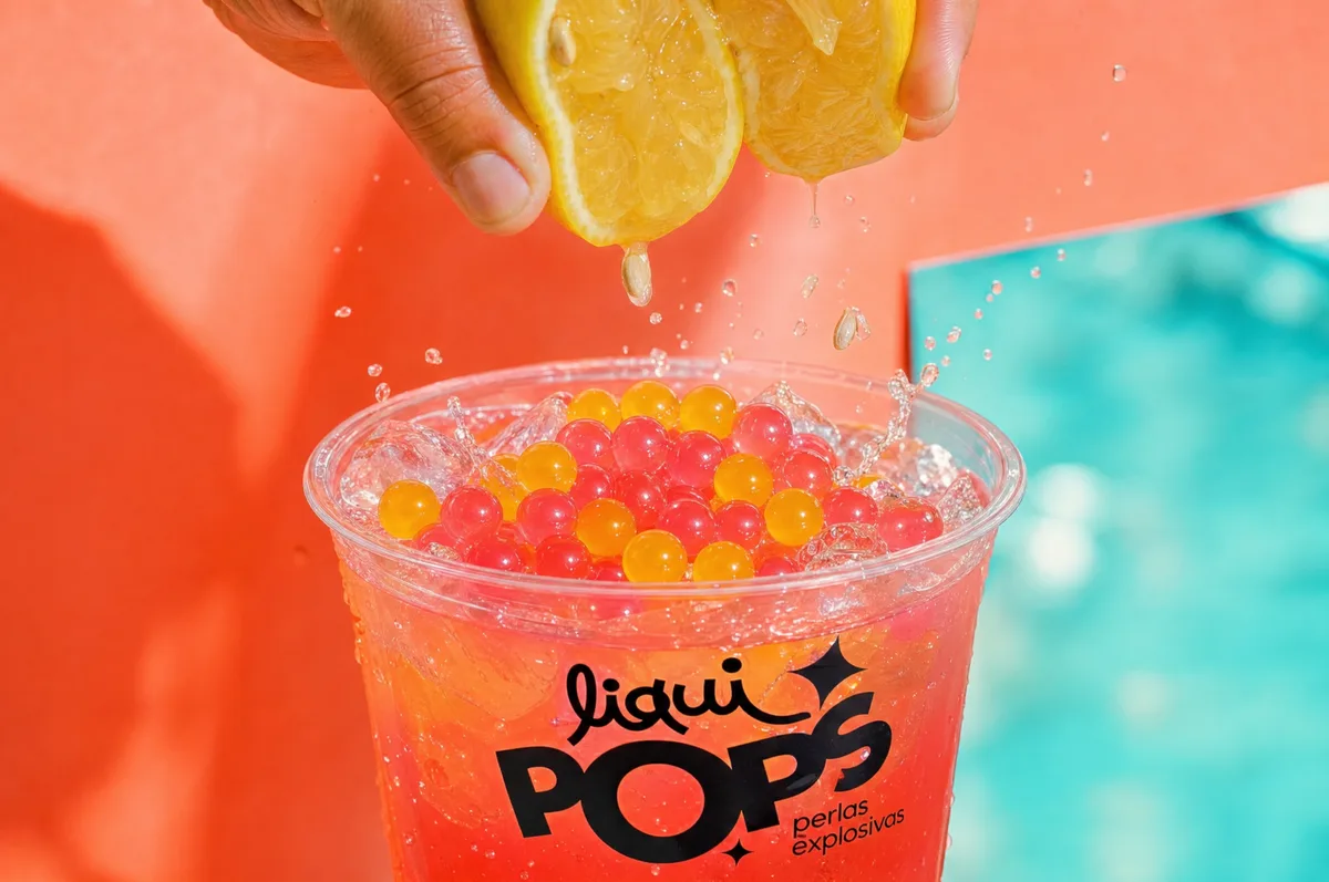 Manos exprimiendo limón sobre un vaso de limonada con perlas explosivas rojas y amarillas LiquiPops — fondo coral y turquesa