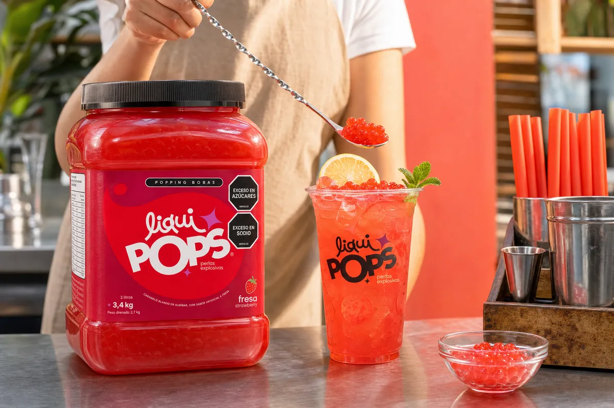 Persona sirviendo con cuchara perlas explosivas rojas de fresa sobre una limonada roja con rodaja de limón y menta en vaso LiquiPops, junto al tarro sabor fresa en la barra