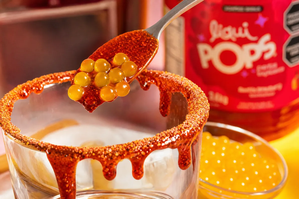 Cuchara con perlas explosivas amarillas sobre el borde escarchado con chamoy de una michelada LiquiPops — primer plano con envase de la marca