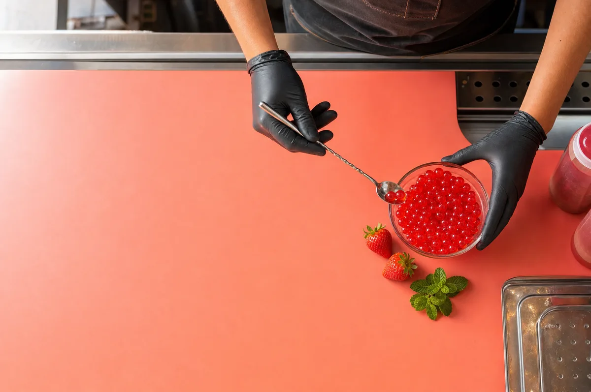Manos con guante sirviendo perlas explosivas rojas de fresa con perlas explosivas LiquiPops desde un bowl con cuchara de bar, fresas y menta sobre superficie coral