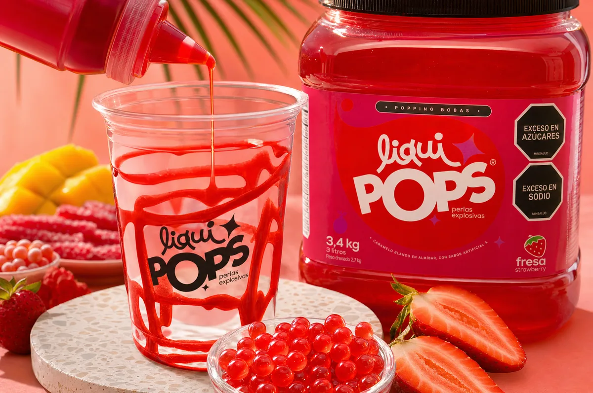 Sirope de fresa decorando un vaso junto a perlas explosivas rojas y el envase sabor fresa LiquiPops — fresas frescas alrededor