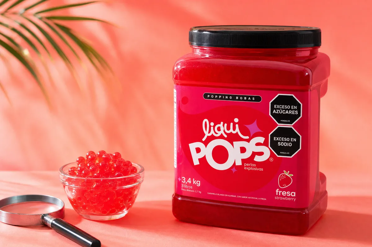 Tarro de perlas explosivas LiquiPops sabor fresa de 3,4 kg junto a un bowl con perlas rojas y una lupa sobre fondo coral con hoja de palma