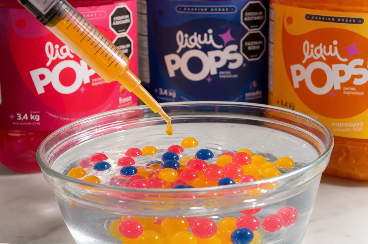 Perlas explosivas rosadas, azules y amarillas formándose en un bowl con jeringa de sirope frente a botes LiquiPops