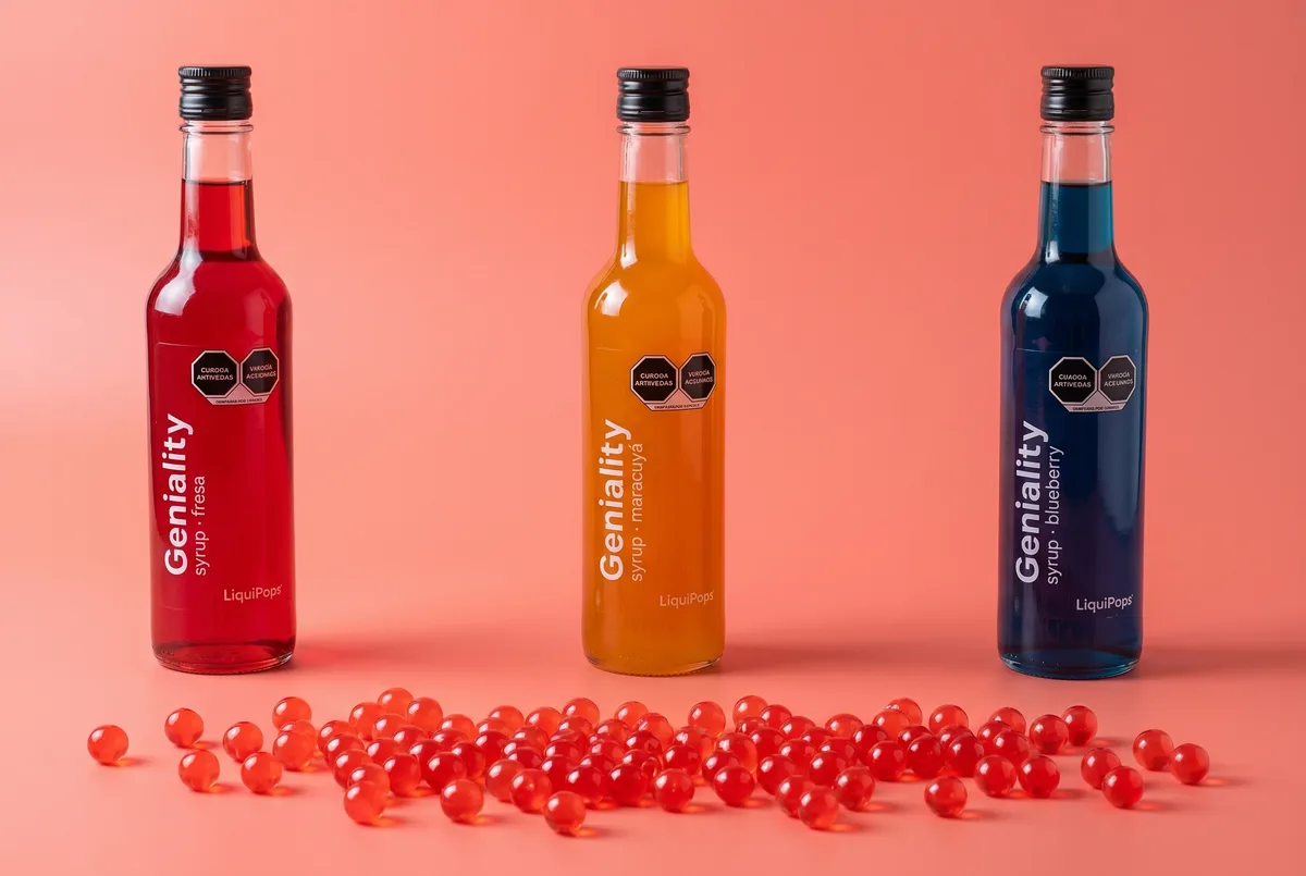 Tres siropes Geniality LiquiPops sabores fresa, maracuyá y blueberry alineados con perlas explosivas rojas regadas sobre fondo coral — bodegón de producto