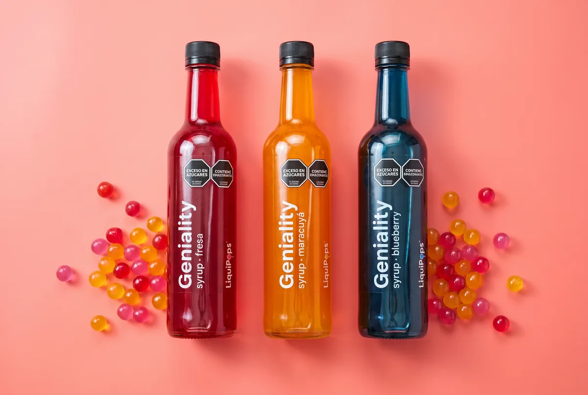 Tres botellas de sirope Geniality by LiquiPops en fresa, maracuyá y blueberry con perlas explosivas LiquiPops alrededor — insumos para bebidas de sabores