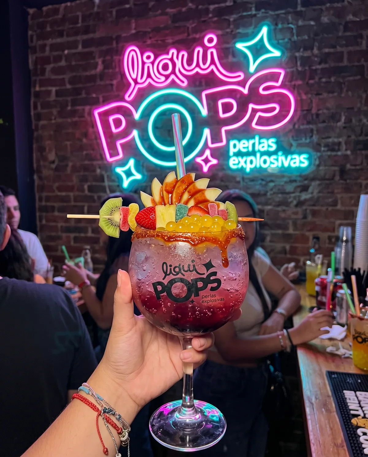 Mano sosteniendo una soda loca de mango con perlas explosivas azules LiquiPops en un bar bajo el letrero neón — pasador de frutas y escarcha de chamoy