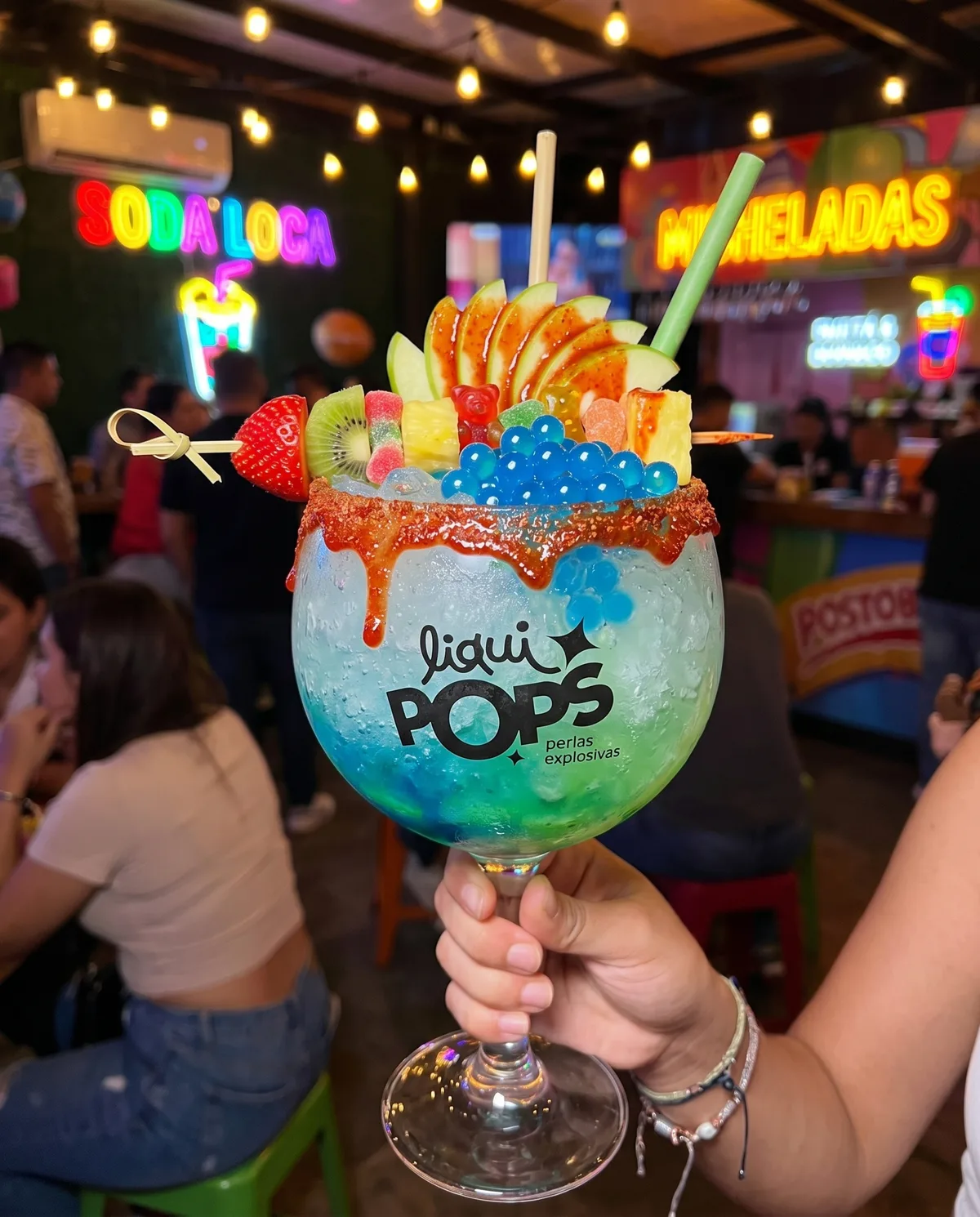 Mano sostiene copa de soda loca con perlas explosivas azules LiquiPops, frutas y chamoy — fondo con neón soda loca y micheladas