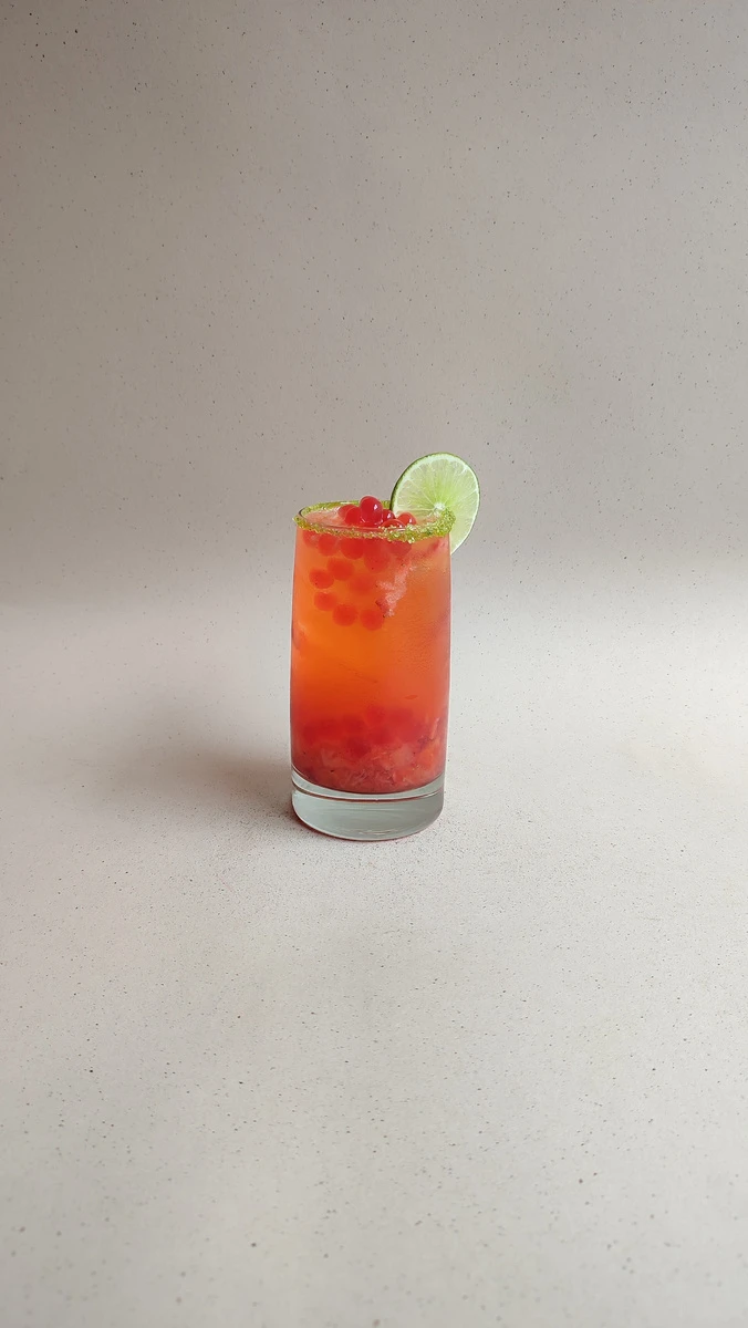 Cóctel de fresa y aguardiente con perlas explosivas LiquiPops, borde escarchado y limón en vaso alto sobre fondo gris