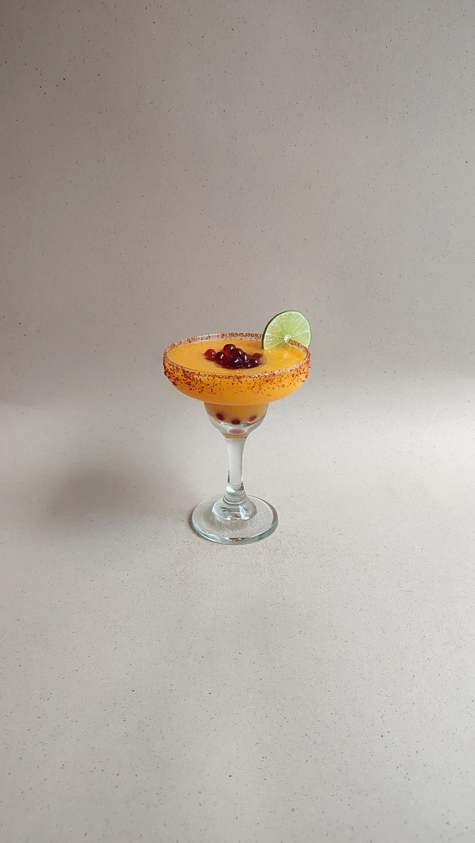 Margarita de mango con perlas explosivas LiquiPops, escarchado de chile y rodaja de limón en copa sobre fondo gris